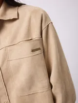 Campera de estilo camisero con efecto gamuzado, color beige, de corte recto y holgado. Presenta cuello clásico, cierre frontal con botones, mangas largas con puños abotonados y bolsillos frontales aplicados.