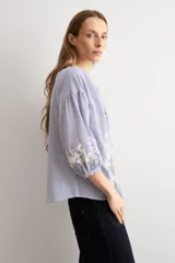 Blusa de manga larga con estampado de rayas verticales en tonos celestes y blancos. Presenta un escote en V con cierre frontal de botones y detalles de bordados florales en color crema en el frente y en los puños.