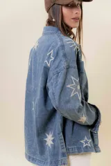 Camisa de denim de lavado medio, con calce relajado y ligeramente oversize. Presenta bordados o estampados de inspiración celestial (soles, lunas y estrellas) en color claro en la parte delantera y mangas. Tiene cuello, bolsillos delanteros con tapa y cierre frontal con botones.