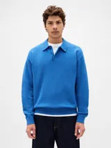 Buzo tipo polo de tejido suave, color azul, con cuello camisero y cierre frontal de dos botones. Presenta puños y ruedo acanalados.
