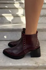 Bota corta de cuero color bordó, con taco cuadrado negro y suela de plataforma. Presenta detalles de tachas metálicas plateadas a lo largo de la costura frontal y lateral, con cierre interno.