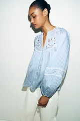 Blusa de rayas verticales blancas y celestes, con cuello redondo y escote en pico. Presenta detalles de bordados perforados y puntillas en el pecho y las mangas. Tiene mangas largas abullonadas con puños elásticos y cierre frontal con botones forrados.