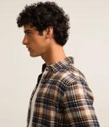 Camisa de flanela con estampado a cuadros en tonos beige, azul y rojo. Presenta cuello clásico, cierre frontal con botones y mangas largas con puños abotonados.