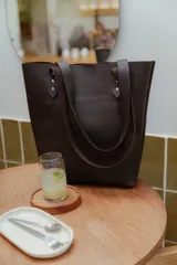 Cartera tipo tote bag de cuero marrón, con diseño minimalista, asas largas y detalles de remaches metálicos en la unión de las asas.