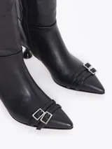 Bota de caña alta confeccionada en cuero vacuno liso, con taco laqueado de altura intermedia y punta fina. Presenta detalles de hebillas metálicas en la caña y en la puntera, incluyendo un dije en forma de corazón.