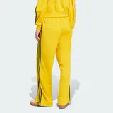 Pantalón deportivo Adidas de corte holgado, color amarillo brillante con las clásicas tres tiras verdes en los laterales y el logo del trifolio bordado en la pierna izquierda. Cuenta con cintura elástica y bolsillos laterales con cierre.