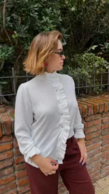 Camisa blanca de textura sutil, con cuello alto tipo victoriano y detalle de volados asimétricos a lo largo de la botonadura frontal. Presenta mangas largas con puños terminados en volados.