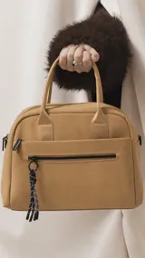 Cartera de mano color beige, confeccionada en tela impermeable, con doble asa corta y correa larga ajustable y desmontable. Presenta cierre principal, bolsillo frontal con cierre y detalle de colgante decorativo.