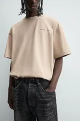 Camiseta de manga corta y cuello redondo, color beige claro, con corte relajado (relaxed fit) y un pequeño texto bordado en el pecho.