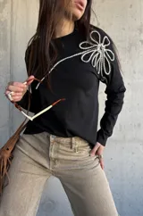 Remera de manga larga color negro, de corte recto, con aplique decorativo en relieve con forma de flor grande en el hombro confeccionado con cordón trenzado en blanco y negro.