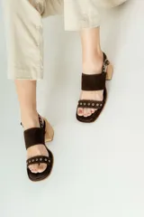 Sandalias de gamuza color marrón con taco cuadrado de madera. Presentan dos tiras anchas, una superior lisa y una inferior decorada con ojales metálicos plateados. Cuentan con cierre de pulsera con hebilla ajustable en el tobillo.