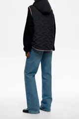 Chaleco acolchado color crema con capucha integrada, cierre frontal de botones a presión y vivos en contraste color negro. Presenta diseño de costuras geométricas y bolsillos laterales.