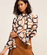 Blusa de chiffon con estampa de eslabones y argollas entrelazadas en tonos marrones y negros sobre fondo blanco. Presenta cuello alto, mangas largas acampanadas y cierre tipo gota en la espalda.