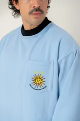 Buzo de cuello redondo color celeste, con bolsillo frontal bordado que presenta el sol de la bandera uruguaya, cuatro estrellas y la frase 'Heroicos sabremos cumplir'.