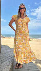 Vestido largo amarillo con estampado floral en tonos rosa, blanco y celeste. Tiene mangas cortas con volados y un volante en el ruedo.
