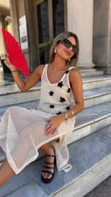 Vestido largo de tejido de red blanco, con apliques de flores negras en relieve. Lleva tirantes finos y tiene un corte recto con abertura lateral. Se usa sobre una prenda interior blanca.