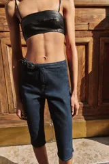 Jeans de tiro medio en denim azul oscuro, con diseño capri de pernera entallada. Presenta cierre frontal con cremallera y botón, y un cinturón integrado con hebilla metálica en la cintura.