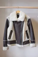 Campera tipo gamulán de cuero genuino color marrón oscuro, con interior y detalles en piel sintética color crema. Presenta cierre frontal cruzado, bolsillos laterales con cierre y puños con borde de piel.