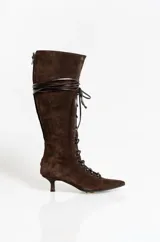 Bota alta de gamuza color marrón oscuro, con diseño de cordones frontales y cierre lateral. Presenta una silueta estilizada con punta fina y un taco bajo tipo kitten heel.