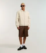 Campera masculina de corte boxy en color beige, con cuello inglés, cierre frontal de cremallera, mangas largas con puños elásticos y ruedo con elástico. Presenta bolsillos laterales y detalles de recortes en el diseño.