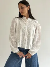 Blusa de brodery color blanco, con cuello redondo, mangas largas abullonadas con puños elásticos y cierre frontal con botones forrados. Presenta detalles de puntillas verticales en el frente.