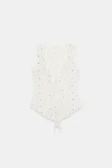 Top sin mangas color blanco roto, con escote en pico y detalle de volantes en los bordes. Presenta pequeñas tachas doradas distribuidas por toda la prenda y cierre frontal con lazadas. El bajo es asimétrico.