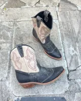 Botas texanas de caña media, confeccionadas en cuero vacuno. Presentan un diseño bicolor que combina el color negro con paneles en color crema, decorados con costuras bordadas en hilo rojo. Cuentan con taco cuadrado de madera y puntera estilizada.