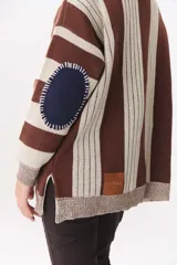 Buzo de punto con diseño de rayas verticales en tonos verde y azul marino. Presenta cuello alto con cierre frontal, hombros caídos y coderas en contraste de color marrón con costuras decorativas visibles.