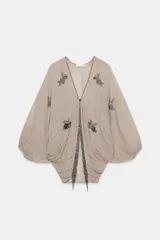 Top de viscosa color beige con escote en V, mangas murciélago abullonadas por debajo del codo y bajo asimétrico. Presenta delicados bordados con aplicaciones de abalorios y cuentas brillantes en el frente y las mangas.