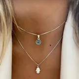 Conjunto de dos collares plateados de cadena fina tipo caja. El collar superior tiene un dije de ojo turco redondo con centro azul oscuro y borde plateado. El collar inferior tiene un dije de mano Hamsa plateada.