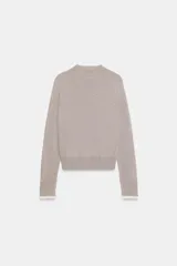 Chaqueta de punto color beige, de manga larga y corte corto, con cuello redondo y puños terminados en puntilla blanca a contraste.