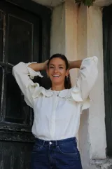 Camisa de color natural con cuello redondo, mangas largas con puños y detalle de volados amplios en la parte superior delantera y hombros. Presenta cierre frontal con botones.