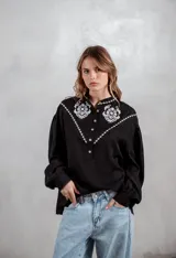 Camisa negra de manga larga con bordados blancos en la parte superior delantera y hombros. Presenta cuello mao, cierre frontal con botones y mangas con puños ajustados.