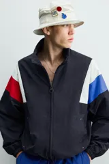 Chaqueta técnica de corte relajado con cuello subido y cierre frontal de cremallera bidireccional. Presenta un diseño color block en tonos azul marino, blanco, rojo y azul eléctrico. Incluye bolsillos frontales de vivo, puños y ruedo elásticos, y forro interior de red.