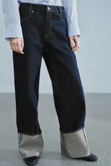 Jeans de tiro bajo y pernera ancha, confeccionados en denim azul oscuro con pespuntes en contraste. Cuentan con un diseño de cinco bolsillos, cierre frontal con cremallera y botón metálico, y destacan por su amplio doblando en el bajo que deja ver el revés de la tela con detalles de costura selvedge.