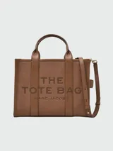 Bolso tipo tote de cuero negro, modelo The Leather Medium Tote Bag de Marc Jacobs. Presenta doble asa superior, correa ajustable para el hombro y el texto grabado "THE TOTE BAG MARC JACOBS" en el frente.