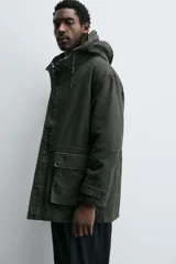Parka color verde militar de corte relaxed fit, confeccionada en tejido de algodón con interior acolchado. Cuenta con cuello subido, capucha ajustable con cordón, cierre frontal de cremallera oculta por solapa con botones a presión y amplios bolsillos frontales de plastrón con solapa.