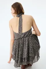 Vestido corto de gasa transparente en color negro con estampado floreado. Presenta escote halter alto, espalda abierta, elástico fruncido en la cintura y falda escalonada con vuelo. Incluye forro interior.
