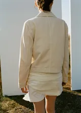 Chaqueta de algodón estilo tejano en color beige, con cuello camisero, manga larga, dos bolsillos de parche en el frente y cierre delantero de botones.