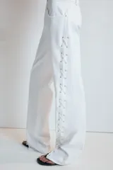Jeans rectos de tiro medio en color blanco, con cinco bolsillos, trabillas y cierre frontal con cremallera. Presenta detalle de cordones cruzados con ojalillos metálicos a lo largo de los laterales de ambas piernas.