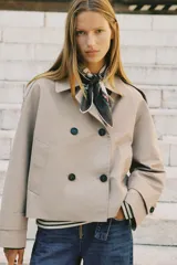 Chaqueta corta estilo trench color marrón oscuro, de corte cruzado con doble botonadura, cuello solapa y hombreras. Presenta mangas largas con puños anchos y bolsillos delanteros de vivo.