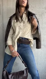 Camisa de color beige con diseño de broderie anglaise, cuello clásico, cierre frontal con botones y mangas largas.