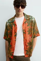 Camisa de corte relajado confeccionada en tejido fluido con mezcla de viscosa y lino. Presenta cuello bowling, manga corta y cierre frontal con botones. Diseño con estampado floral en tonos verdes y naranjas.
