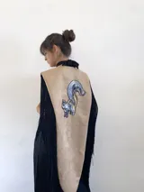 Poncho estructurado color beige con diseño asimétrico, más corto en el frente y largo en la parte posterior. Presenta un bordado de ardilla con lentejuelas en la espalda y flecos largos negros que rodean toda la prenda.