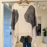 Blusa blanca de corte holgado y diseño asimétrico, confeccionada en tela translúcida con estampa de hojas en tono gris oscuro y mangas murciélago.