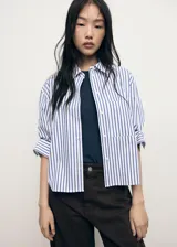 Camisa crop de corte recto confeccionada en mezcla de algodón, poliéster y viscosa. Presenta diseño de rayas verticales en azul y blanco, cuello camisero, bolsillo aplicado en el pecho y cierre delantero con botones.