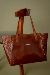 Bolso tipo shopper de cuero color marrón, con laterales texturizados efecto cocodrilo y detalles de costuras decorativas en color claro. Cuenta con doble asa de hombro y placa metálica con logo en el frente.