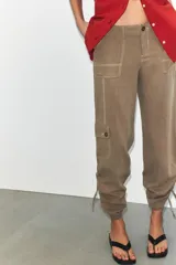 Pantalón de tiro medio confeccionado en tejido fluido color tostado. Presenta bolsillos de plastrón en el delantero y en la pierna, solapas en la espalda y detalle de frunces ajustables en el bajo. Cierre frontal con cremallera y botón.