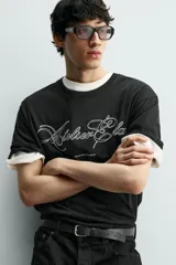 Camiseta de corte relajado color negro, con cuello redondo y manga corta. Presenta un bordado frontal con texto en color blanco a contraste.