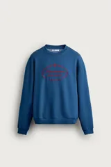 Sudadera azul con corte relaxed fit, cuello redondo y manga larga, confeccionada en tejido de algodón. Presenta un bordado frontal a contraste en color rojo con diseño ornamental y texto.